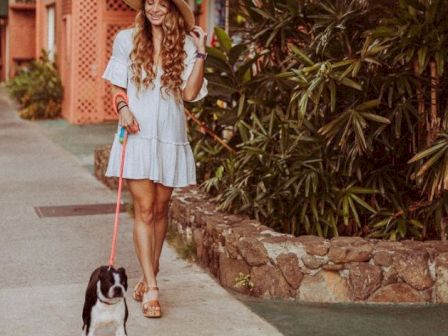 A woman in a hat and dress is walking a small black and white dog on a leash near greenery and buildings.