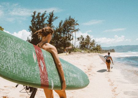 Two people are walking on a sandy beach carrying surfboards. One person leads while the other follows. The sky is clear with a few clouds.