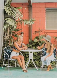 Two people sit at a table outside near surfboards and plants, against a red wall with windows and a door.