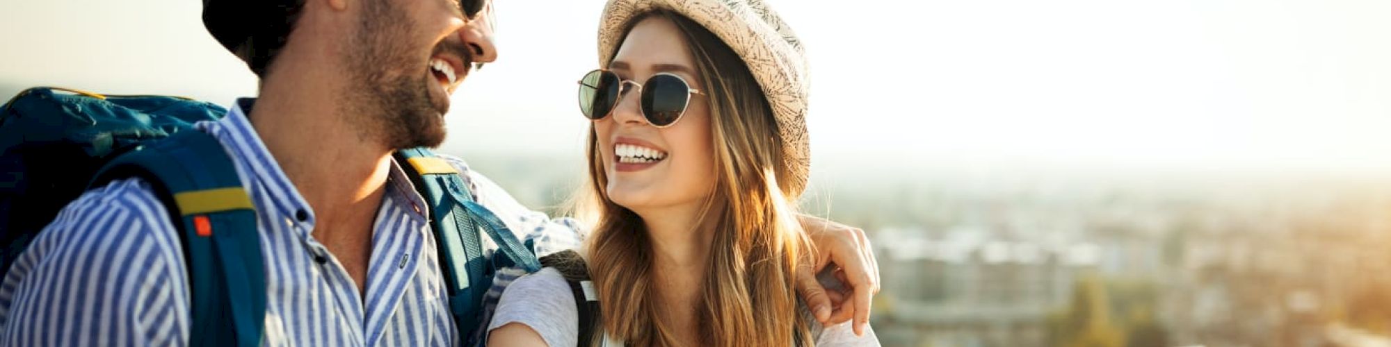A smiling couple wearing sunglasses and hats, with backpacks, enjoying an outdoor setting while holding a map, suggesting they are traveling.