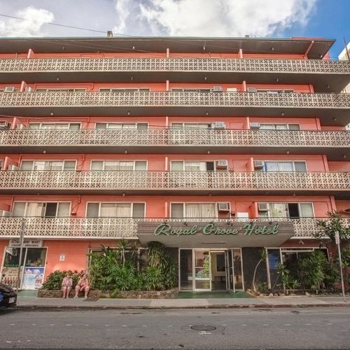This image shows a multi-story building with a sign reading "Royal Grove Hotel" on the front, surrounded by plants and parked cars.