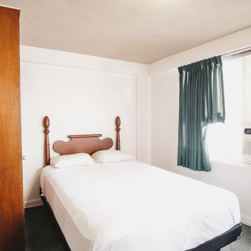 A small bedroom with a bed, wooden headboard, nightstand, closet, green curtains, and a window air conditioner.