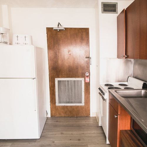 The image shows a small kitchen with a white refrigerator, wooden cabinets, a stovetop, a sink, and a wooden door with a grill, likely leading outside.