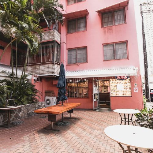 The image shows an outdoor seating area with a few tables, shaded by an umbrella, next to a pink building.