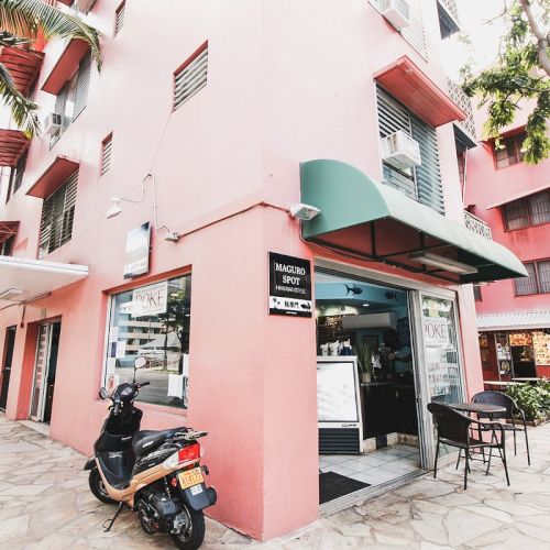 Image of a street corner with a pink building, a parked scooter, outdoor tables and chairs, and a business with a green awning.