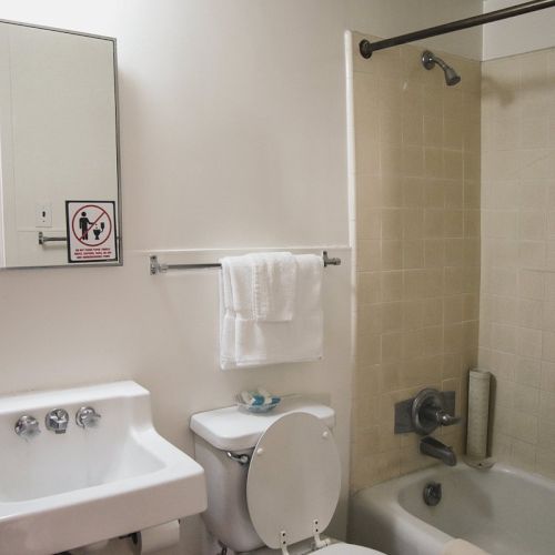 This image shows a bathroom with a mirror, sink, toilet, and bathtub, all in an off-white color scheme. Towels are hung above the toilet.