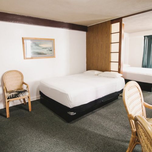 The image shows a hotel room with two beds, wicker chairs, a microwave, and a framed picture on the wall, separated by partition doors.