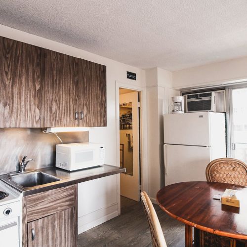 This image shows a small kitchen with wooden cabinets, a stove, a microwave, a fridge, a sink, a round table with chairs, and a window.