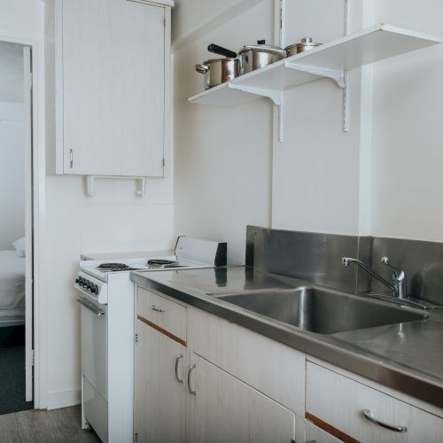 A compact kitchen with white cabinets, stainless steel countertops, and a small stove, adjacent to a bedroom visible through an open doorway.