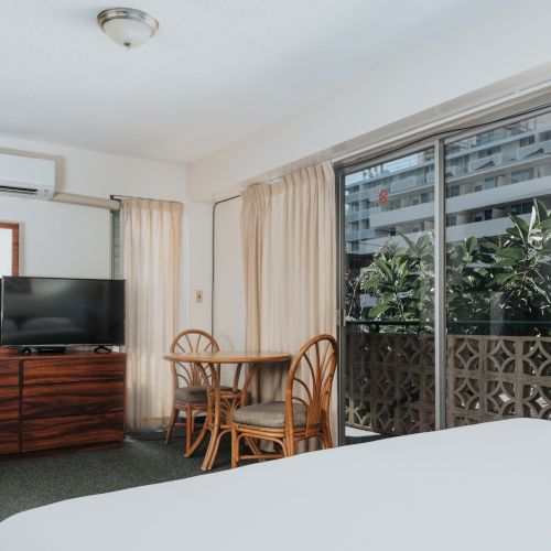 A hotel room with a bed, TV, table with chairs, dresser, sliding glass door, and air conditioner, with curtains and a view of buildings and plants.