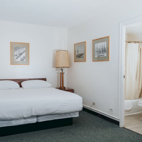 A hotel room with a bed, two lamps, three framed pictures on the wall, and an adjoining bathroom with a shower curtain and toilet.