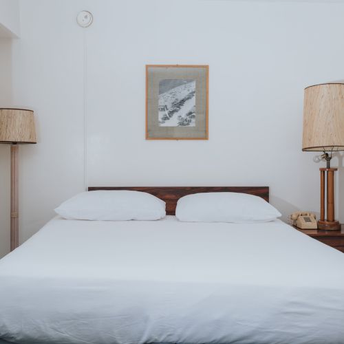 The image shows a simple bedroom with a white bed, wooden headboard, two lamps on either side, and framed pictures on the walls.