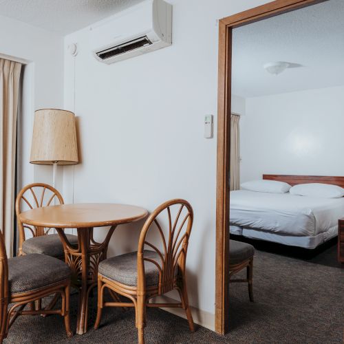 The image shows a small dining area with a round table and four chairs, next to a doorway leading to a bedroom with a double bed.
