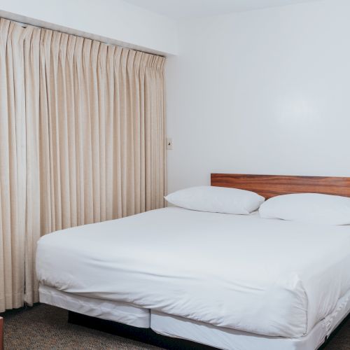A neatly made double bed in a small room with beige curtains and wooden furniture is shown, including bedside tables and a lamp.