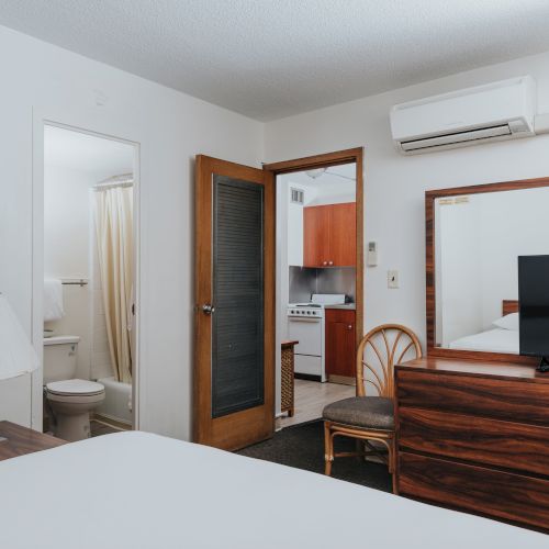 A bedroom features a bed, TV on a dresser, a mirror, a chair, a lamp, a bathroom, and a partially open door to the kitchenette.
