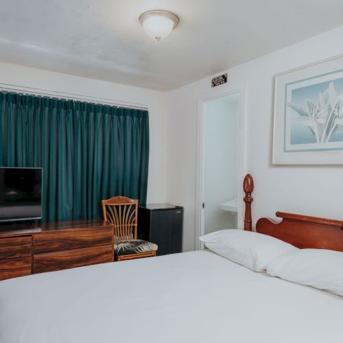 A bedroom with a bed, wooden headboard, TV, dresser, chair, closed curtain, and framed artwork on the wall, in a simple setting.