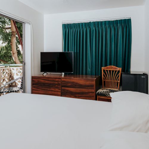 A bedroom with a white bed, a wooden dresser with a TV, a chair, a green curtain, a balcony with tree view, and a small refrigerator.