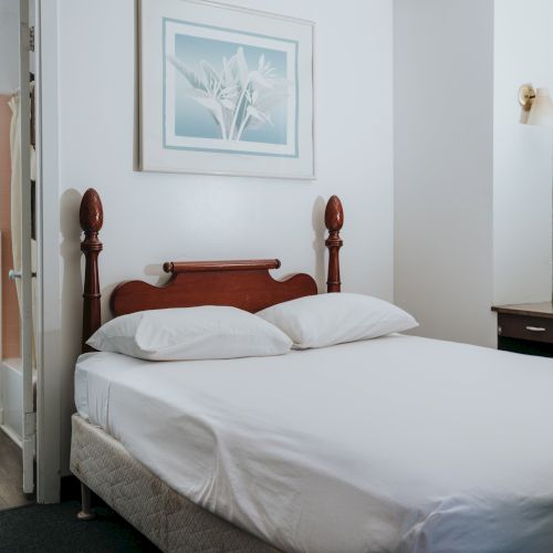 A small bedroom with a double bed, adjacent bathroom, wooden headboard, dresser with mirror, framed picture, and a window with a green curtain, ending the sentence.