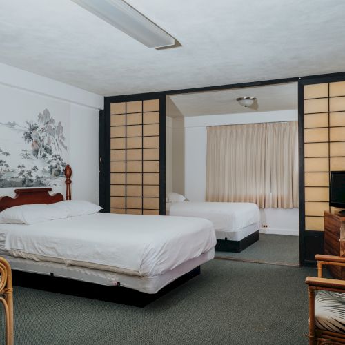 The image shows a spacious bedroom with two beds, a TV, wicker chairs, a wooden desk, and a room divider. There is also a wall mural and carpeted floor.