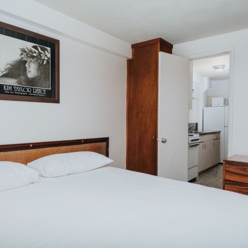 A small, clean bedroom with a double bed, wooden furniture, a TV, and a framed wall art, connected to a kitchen through an open door ending the sentence.