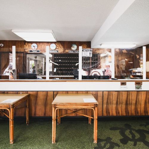A reception desk with clocks, a key rack, and two wooden tables in front. The room has wooden panel walls and green carpet flooring.