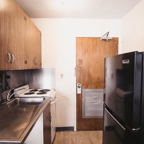 A small kitchen with wooden cabinets, a black refrigerator, an electric stove, a sink, a microwave, and a coffee maker on top of the fridge.