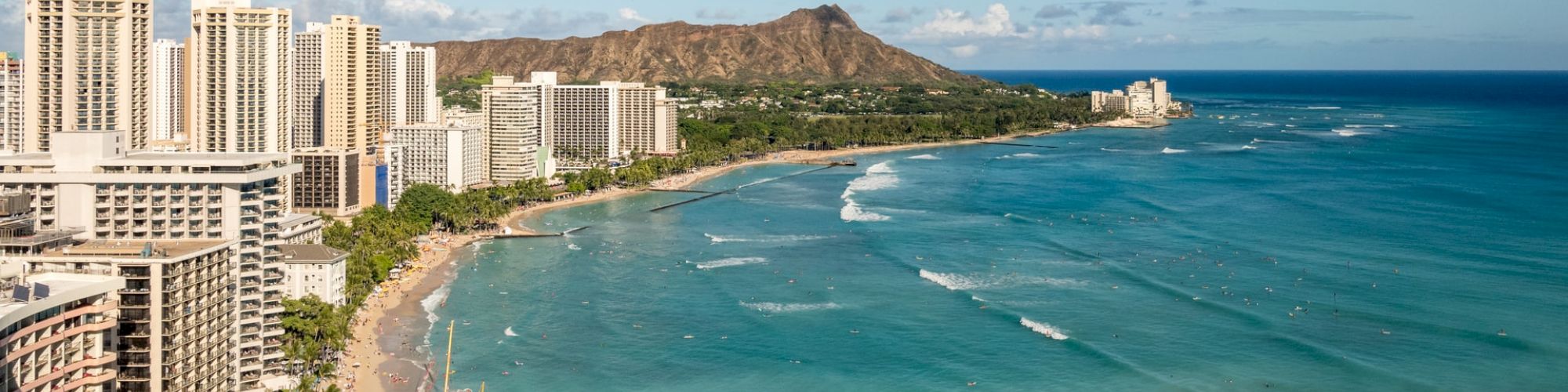 The image shows a coastal cityscape with tall buildings, a sandy beach, and a scenic mountain in the background by a turquoise ocean.
