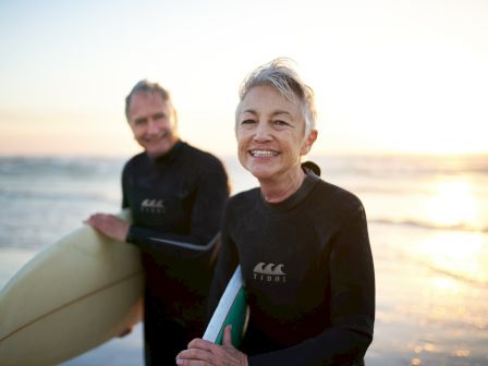Two people in wetsuits, holding surfboards, standing on a beach with the ocean and sunset in the background, looking at the camera.