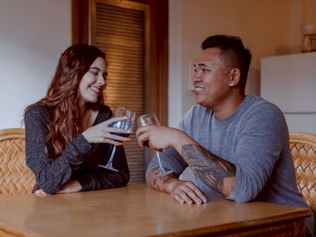 Two people sitting at a table, smiling and clinking their glasses, appearing to be enjoying each other's company.