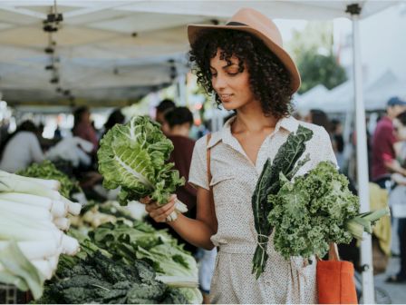 A woman is at a farmer's market, holding fresh leafy greens and wearing a hat. She is surrounded by various vegetables displayed on the stall tables.