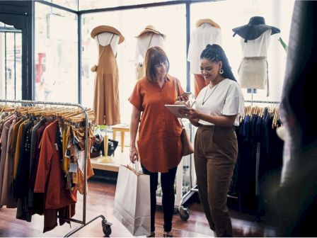 Two women in a clothing store, one holding a shopping bag and the other showing a tablet, surrounded by clothes and mannequins.