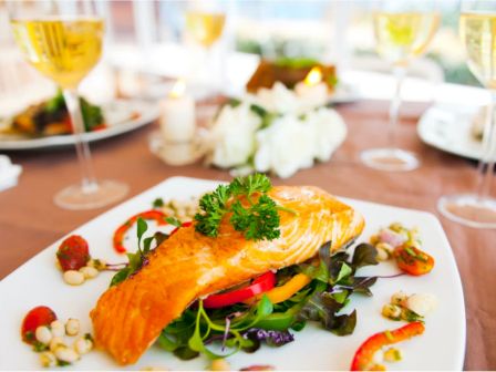A plated dish featuring grilled salmon on a bed of mixed greens with garnishes, surrounded by glasses of white wine on a set dining table.