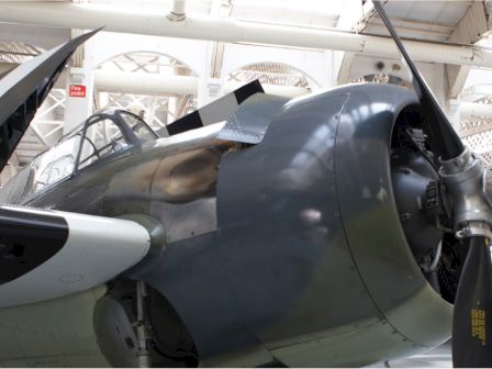 The image shows a close-up view of a vintage aircraft, focusing on its cockpit and propeller, inside a museum or hangar.