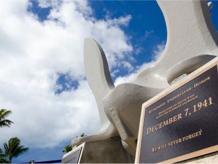 The image shows a memorial plaque dated December 7, 1941, with the inscription "WE WILL NEVER FORGET," against a background of a partly cloudy sky.