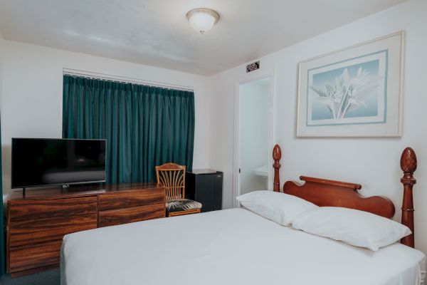 A simple bedroom with a bed, teal curtains, a wooden dresser with a TV, a chair, and a framed picture on the wall ends the sentence.