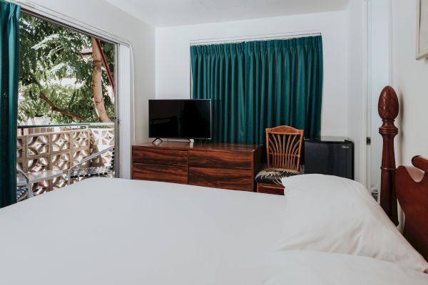 A cozy bedroom with a large bed, TV on a wooden dresser, a chair, green curtains, and a balcony with an outdoor view.
