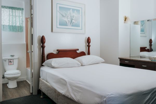 A small bedroom with a bed, pillows, framed artwork, a dresser with a mirror, and an open door revealing a bathroom with a toilet.