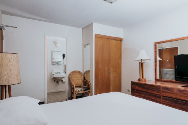 A neatly arranged hotel room with a bed, wooden furniture, a TV, a lamp, and a mirror. The bathroom is visible, with wicker chairs nearby.