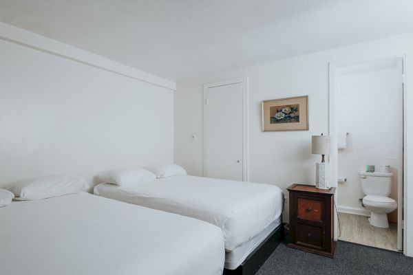 The image shows a clean and simple hotel room with two single beds, a bedside table with a lamp, a painting on the wall, and an adjacent bathroom.