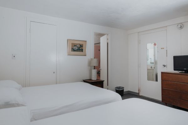 A simple hotel room with two beds, a dresser with a TV, a nightstand with a lamp, and a door with a mirror. A painting is hung on the wall.