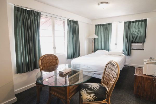 This image shows a cozy bedroom with a bed, green curtains, glass table with chairs, a wooden nightstand, and an air conditioner, all simply decorated.