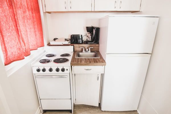 A compact kitchen with a fridge, stove, sink, toaster, coffee maker, white cabinets, and red curtains on a window.
