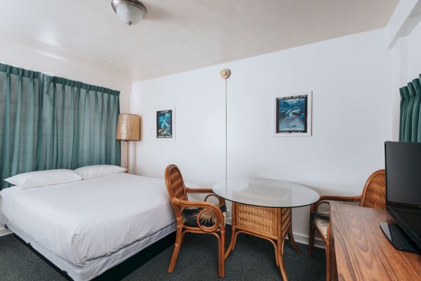 The image shows a hotel room with a bed, two wicker chairs, a glass-topped table, a TV on a wooden dresser, green curtains, and wall art.