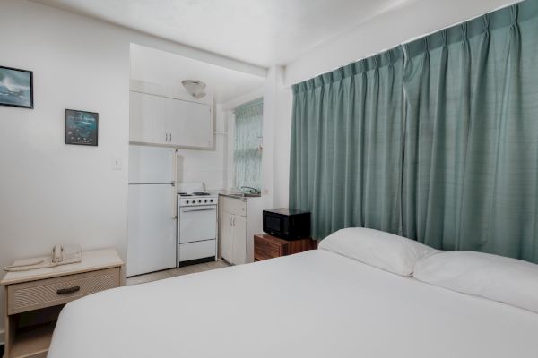 A small room features a bed, a kitchenette with fridge and stove, a microwave, green curtains, and two framed pictures on the wall, ending the sentence.