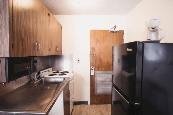 A small kitchen with wooden cabinets, a black refrigerator, a stove, a microwave, and a coffee maker on top of the fridge near a wooden door.