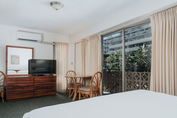 A hotel room with a bed, dresser with a TV, a small table with chairs, and a window with curtains leading to a balcony.