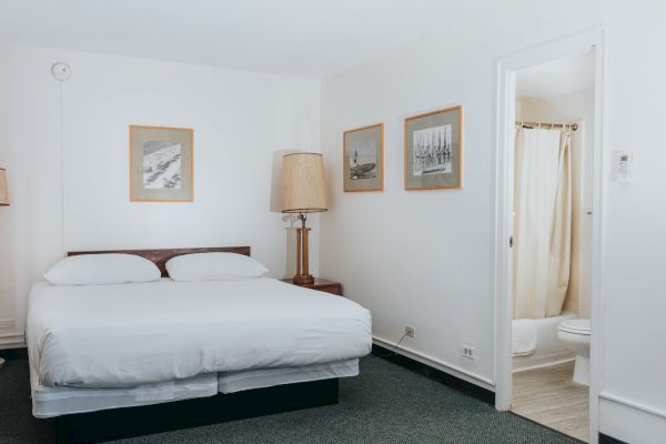 A small, minimalist bedroom with a double bed, bedside table and lamp, three framed pictures, and an open door leading to a bathroom.