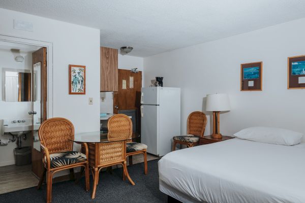 A hotel room with a bed, refrigerator, table with two chairs, a bathroom sink, two framed pictures, and a lamp on a side table.