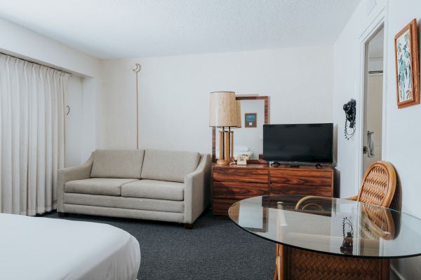 A hotel room features a sofa, a TV on a wooden dresser, a glass table with wicker chairs, and a bed partially visible on the left end of the sentence.