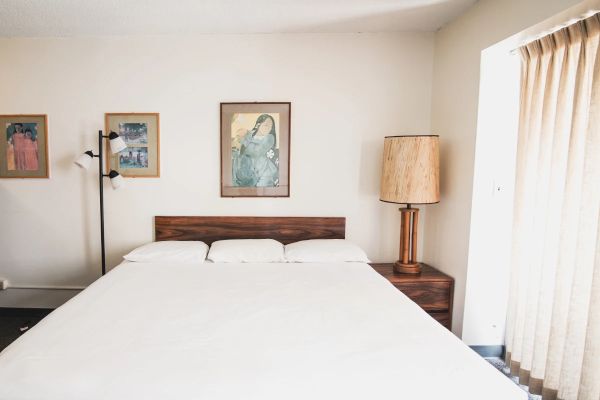 A bedroom with a large bed, three framed artworks on the wall, a floor lamp, a lamp on a nightstand, and a window with curtains ending the sentence.